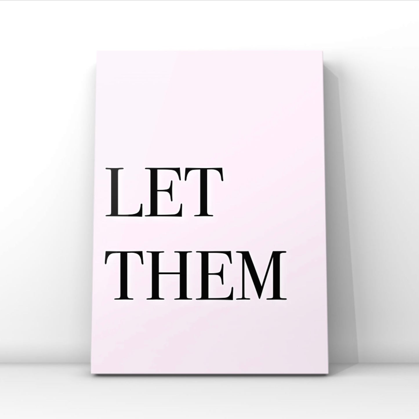 High-impact minimalist pink art print reading "Let Them" in a bold black serif font, leaning against a crisp white gallery wall.