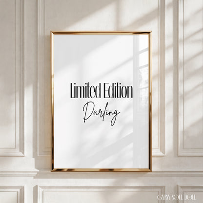 A framed typographic art print with the words 'Limited Edition Darling' in black script and serif text on a white background, set in a gold frame and leaning against a white panelled wall with soft window shadows by Gypsy Soul Doll.