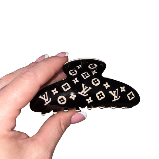 A black hair claw clip with a white  Louis Vuitton monogram pattern, held by a hand with light pink French-tipped nails against a plain white background.