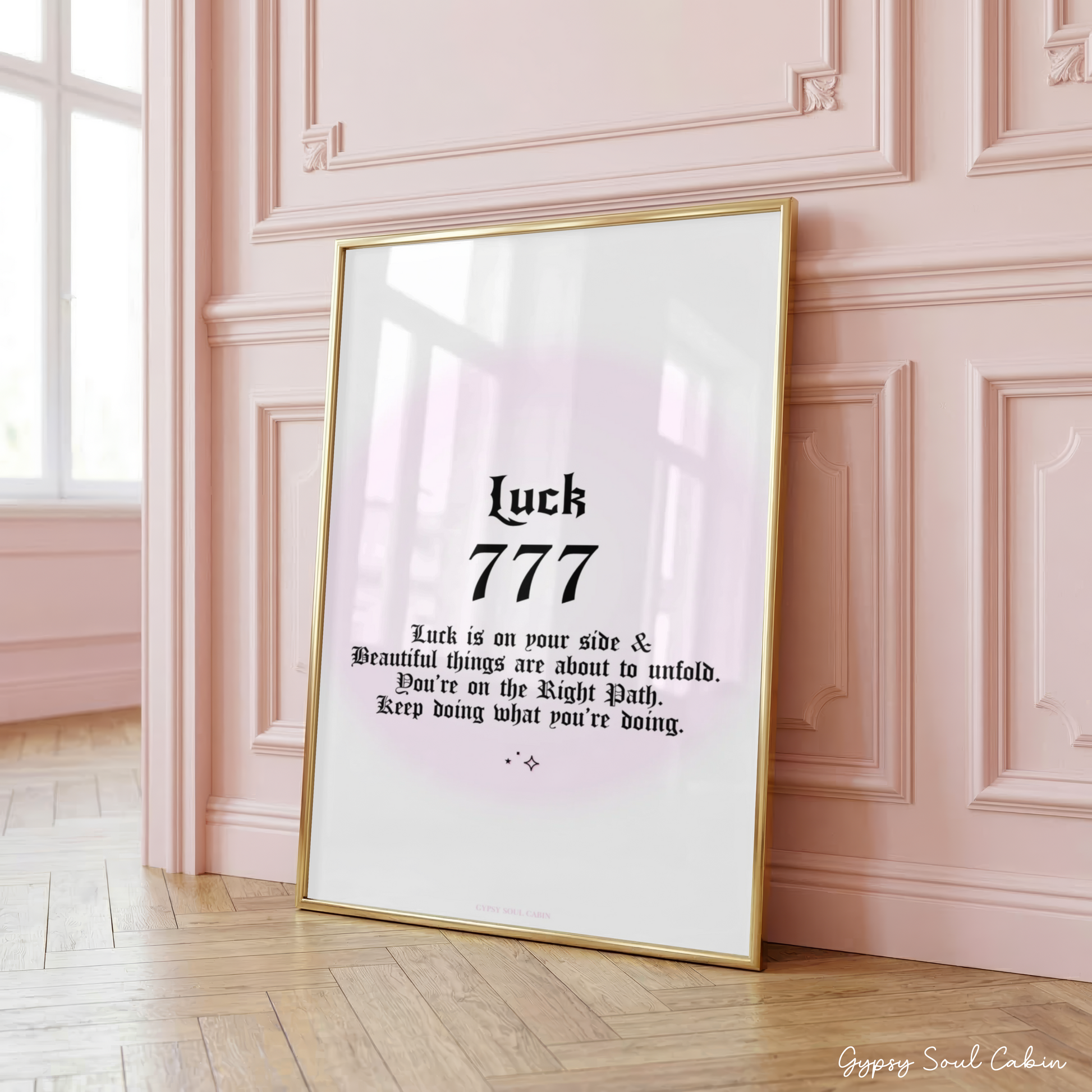 Minimalist A4 manifestation wall art print featuring the word Luck and 777 angel number in bold black gothic typography over a soft pastel pink aura gradient backdrop, framed on a wooden floor against a pink panelled wall.