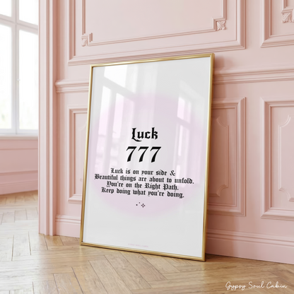 Minimalist A4 manifestation wall art print featuring the word Luck and 777 angel number in bold black gothic typography over a soft pastel pink aura gradient backdrop, framed on a wooden floor against a pink panelled wall.