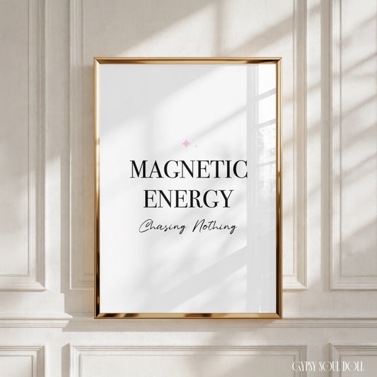 A framed wall art print with the text 'MAGNETIC ENERGY Chasing Nothing' in black lettering on a white background, enclosed in a thin gold frame and mounted on a white panelled wall with soft window shadows by Gypsy Soul Doll.