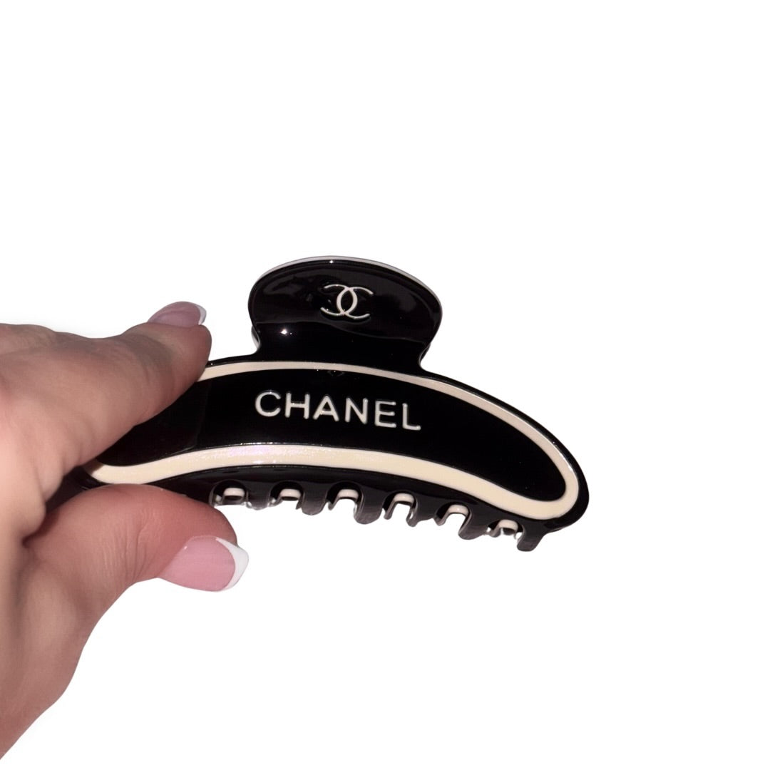 A hand with a classic french manicure holding a curved jet-black hair claw clip featuring cream designer block text and an interlocking circle logo design against a white background.
