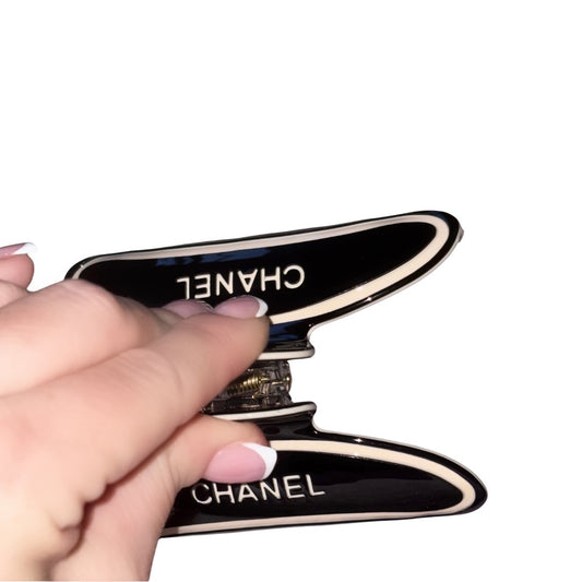 A hand with a classic french manicure holding a curved jet-black hair claw clip featuring cream designer block text and an interlocking circle logo design against a white background.