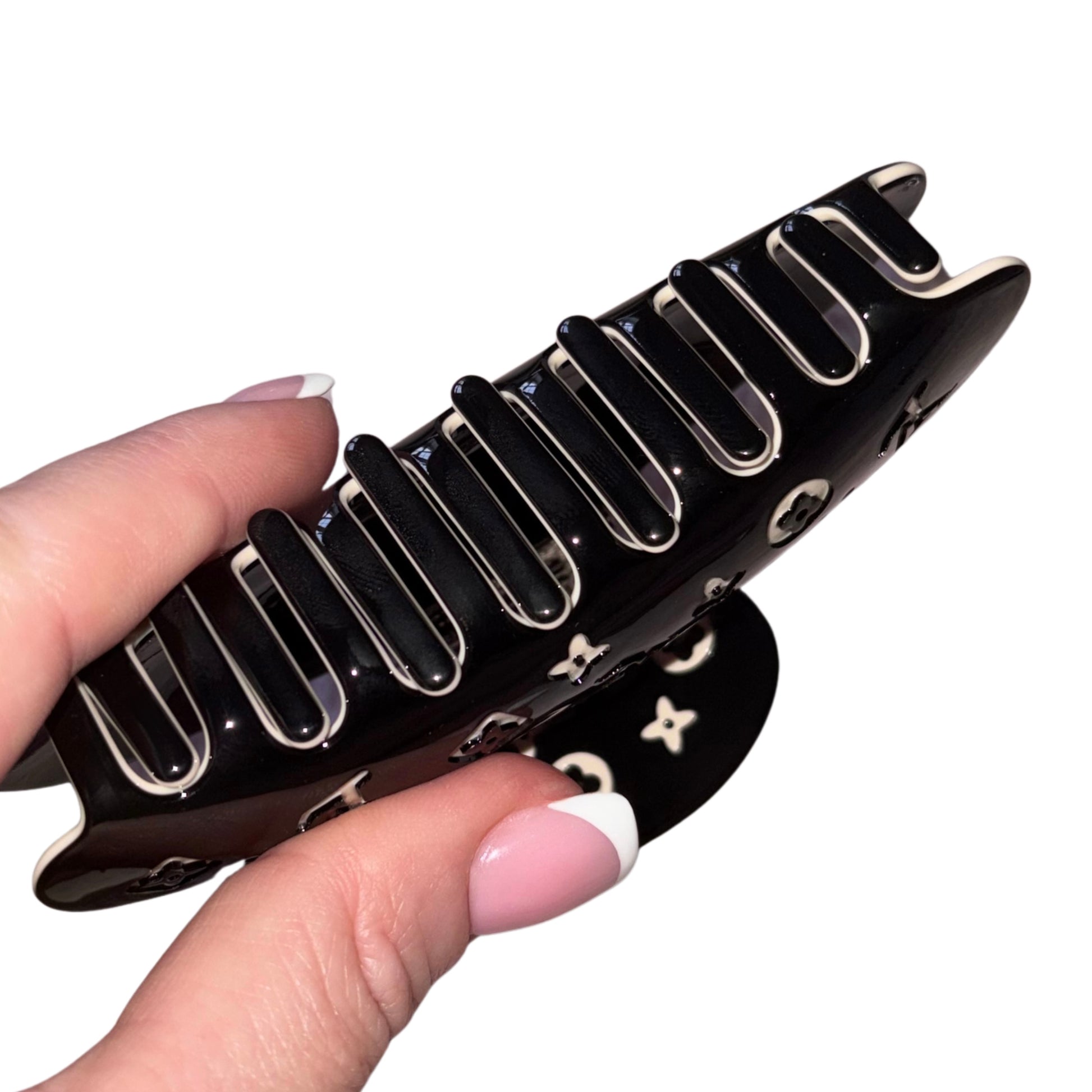 Close-up of a hand with a pink French manicure holding a high-gloss black claw clip hair accessory covered in a cream-white monogram flower print pattern.