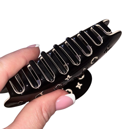 Close-up of a hand with a pink French manicure holding a high-gloss black claw clip hair accessory covered in a cream-white monogram flower print pattern.