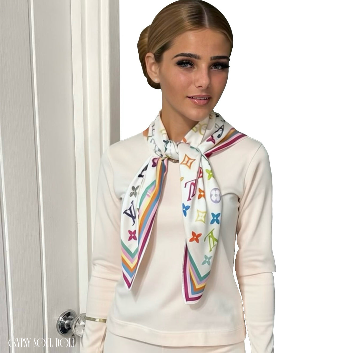A model wearing a cream loungewear outfit, styled with a white scarf featuring a colourful, multicoloured monogram pattern and striped borders. The scarf displays the Louis Vuitton monogram motif.