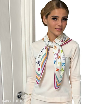 A model wearing a cream loungewear outfit, styled with a white scarf featuring a colourful, multicoloured monogram pattern and striped borders. The scarf displays the Louis Vuitton monogram motif.