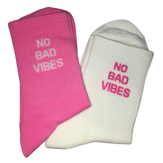 Two folded crew socks on a plain white background, one bright pink and one off-white, each with the phrase 'NO BAD VIBES' embroidered in the opposite color.