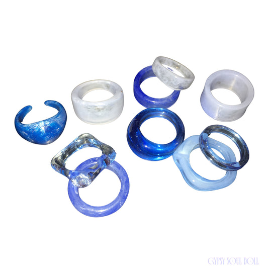 Studio flat lay of the Gypsy Soul Doll limited edition set of 10 retro resin rings featuring cobalt blue dome bands, clear acrylic squares, and marbled white styles against a plain white background with a blue logo.