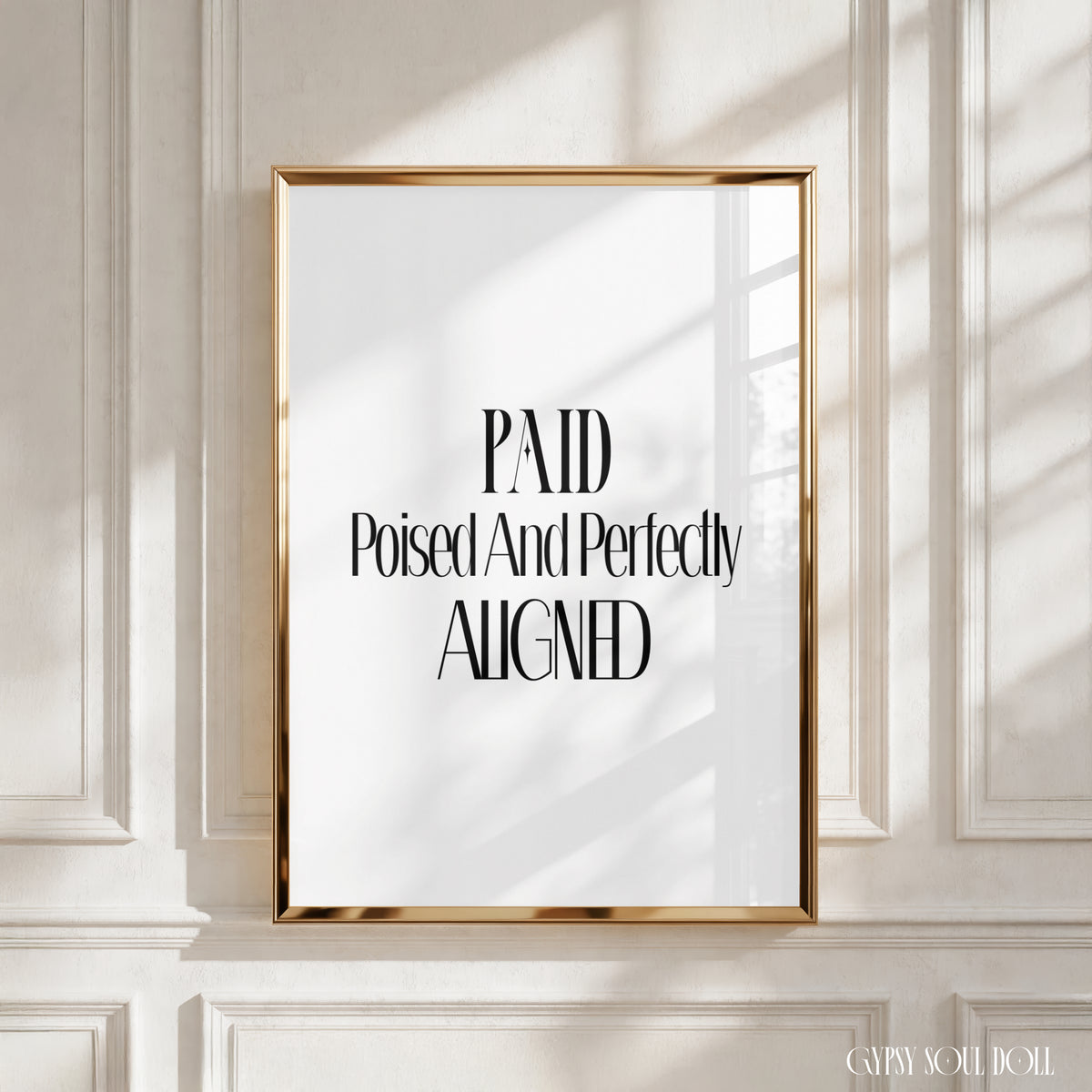 A framed typographic wall art print with the text 'PAID Poised And Perfectly ALIGNED' in black serif lettering on a white background, set in a thin gold frame against a white panelled wall with soft window shadows by Gypsy Soul Doll.