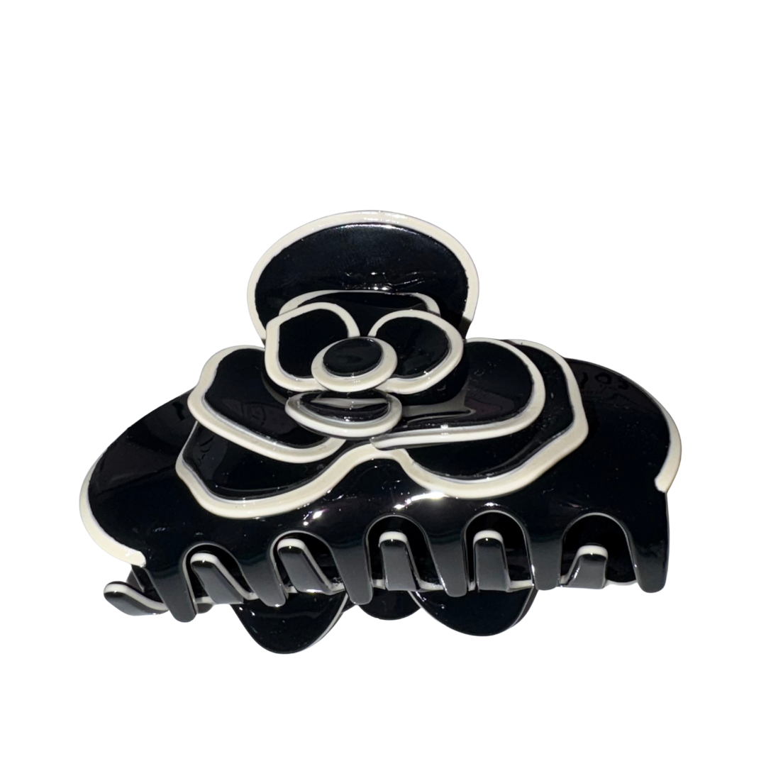 Studio product view of a glossy black acetate flower claw clip featuring an intricate layered camellia blossom design with crisp white contrast piped borders against a white backdrop.