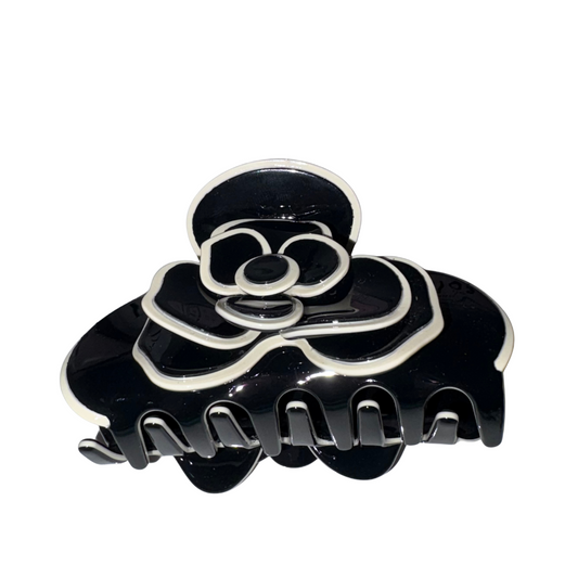 Studio product view of a glossy black acetate flower claw clip featuring an intricate layered camellia blossom design with crisp white contrast piped borders against a white backdrop.
