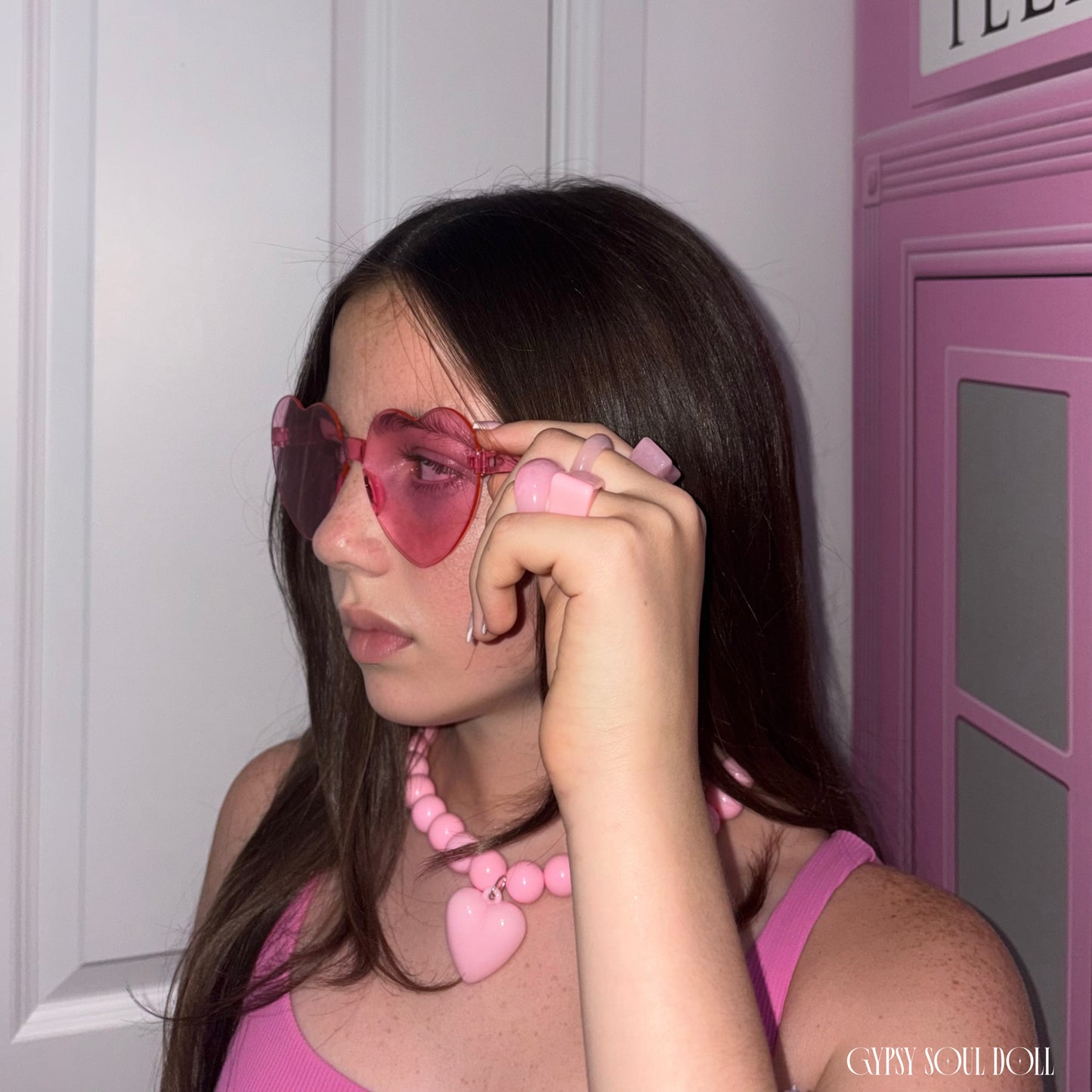 A young model with long dark brown hair wearing pink heart-shaped sunglasses, a pink beaded necklace with a heart pendant, and a pink top, posing next to a pink telephone booth.