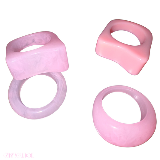 A flat lay studio image displaying a collection of four chunky Y2K style acrylic rings in shades of pastel and translucent pink on a clean white backdrop.