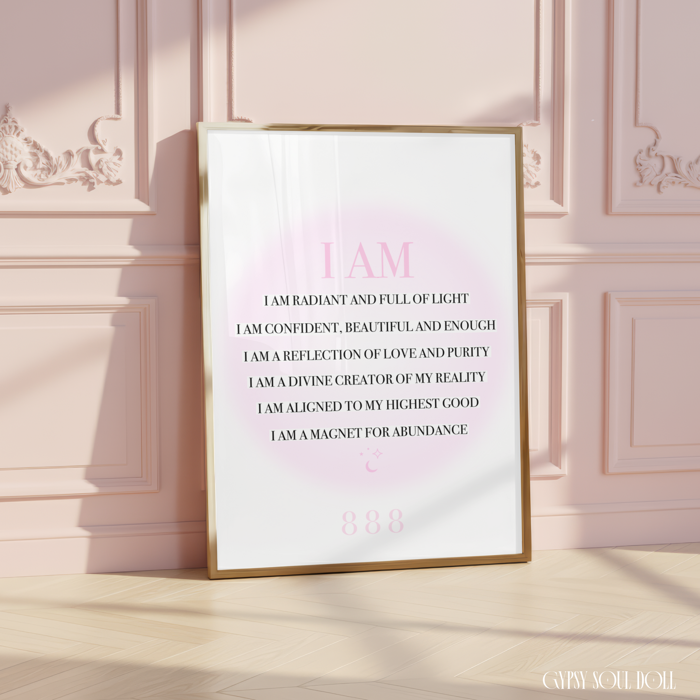 A framed inspirational quote print leaning against a pink paneled wall on a light wood floor. The print has a gold frame, a white background, and pink text that reads, 'I AM I AM RADIANT AND FULL OF LIGHT I AM CONFIDENT, BEAUTIFUL AND ENOUGH I AM A REFLECTION OF LOVE AND PURITY I AM A DIVINE CREATOR OF MY REALITY I AM ALIGNED TO MY HIGHEST GOOD I AM A MAGNET FOR ABUNDANCE.