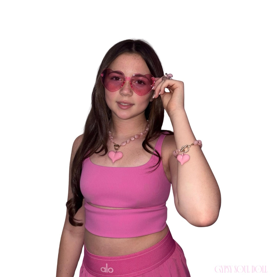 A young woman wearing a matching pink activewear set from alo, featuring a crop top and high-waisted shorts, along with pink heart-shaped sunglasses, a pink heart necklace, and a pink heart bracelet.