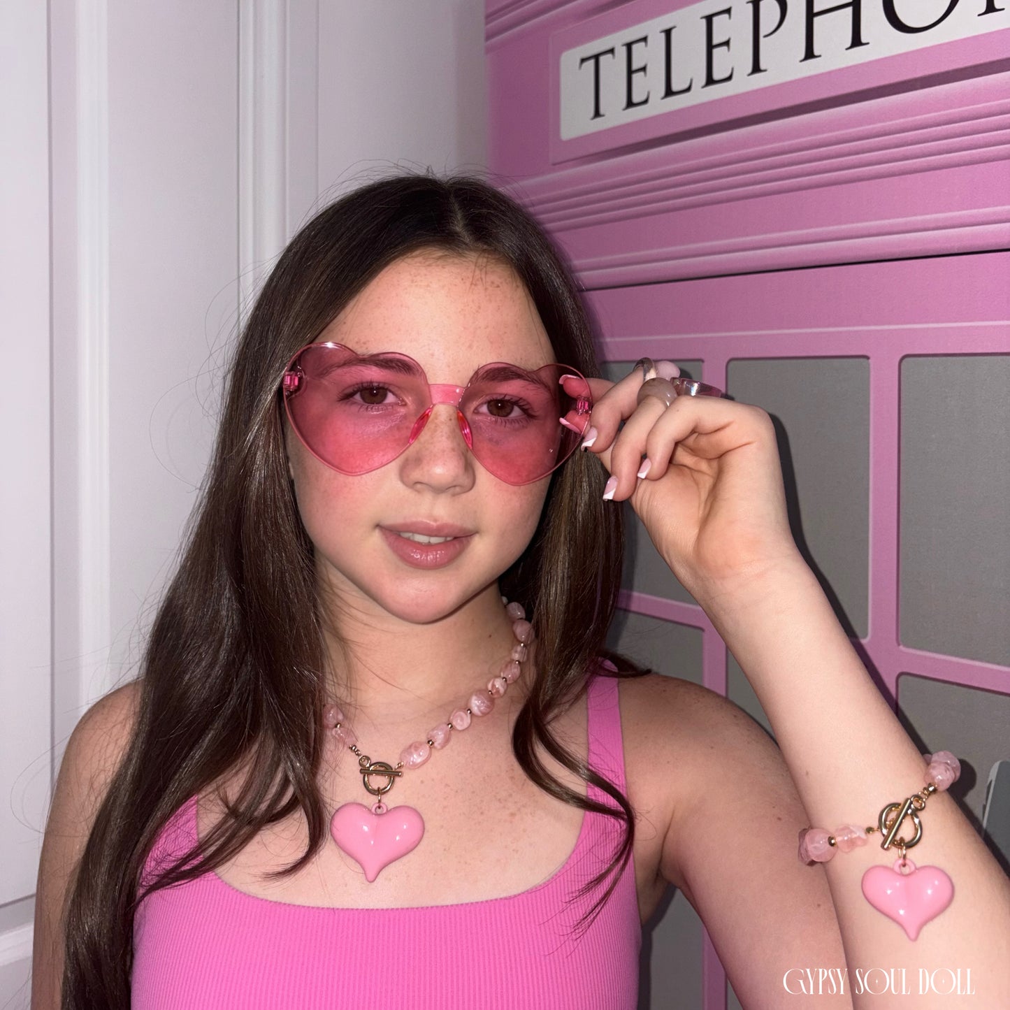 A young model with long brown hair wearing pink heart-shaped sunglasses, a pink beaded necklace with a pink heart pendant, and a pink bracelet with a heart charm by Gypsy Soul Doll posing in front of a pink telephone booth.