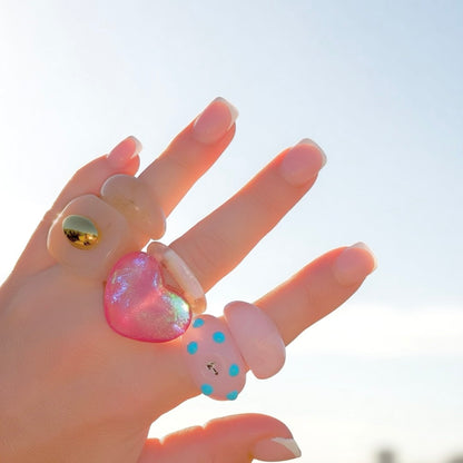 Close-up of a hand with a classic French manicure showcasing a stacked collection of translucent lemon yellow, blue polka dot, and glittery pink heart chunky acrylic statement rings.