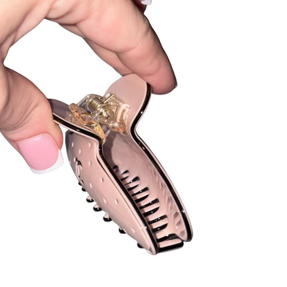 A hand with pink French-tip nails holds a glossy blush pink Chanel hair claw clip with a clear plastic inner section and gold-toned metal hardware against a plain white background.