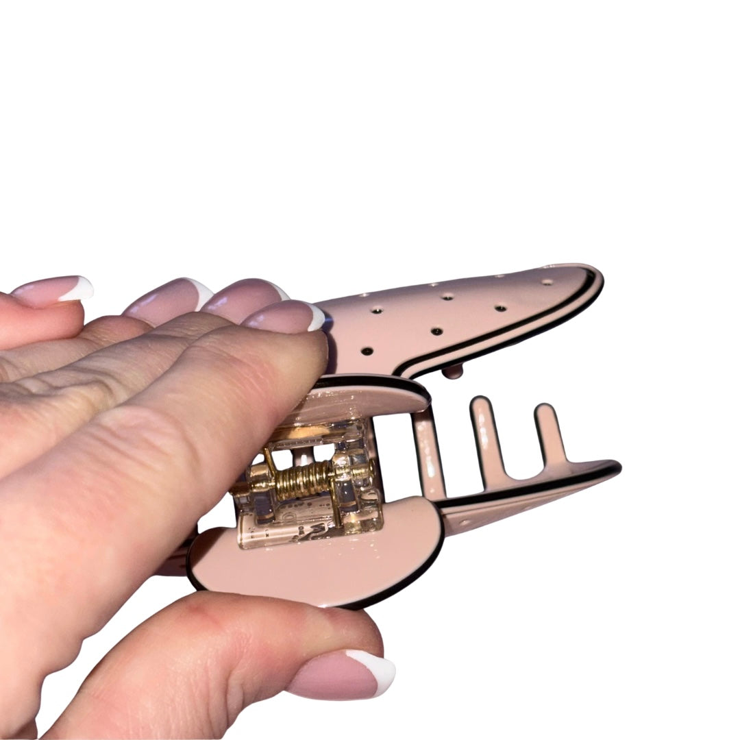 A close-up of a hand holding a rose gold Chanel claw hair clip with a clear plastic spring mechanism, set against a plain white background.