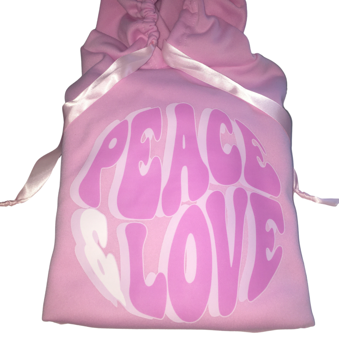 Flat lay close-up of a pink one-size polyester hoodie highlighting the light pink satin ribbon tie strings and large circular pink Peace Love bubble graphic text across the front.