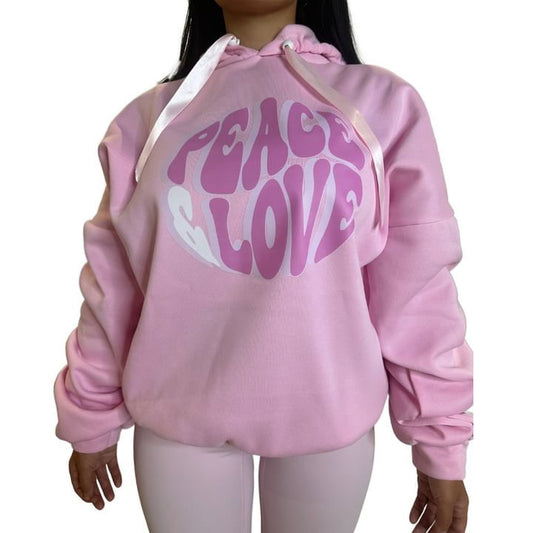 Front view of a model wearing a pink one-size hoodie with scrunched sleeve details, white satin ribbon ties, and a prominent pink Peace Love circular chest graphic.