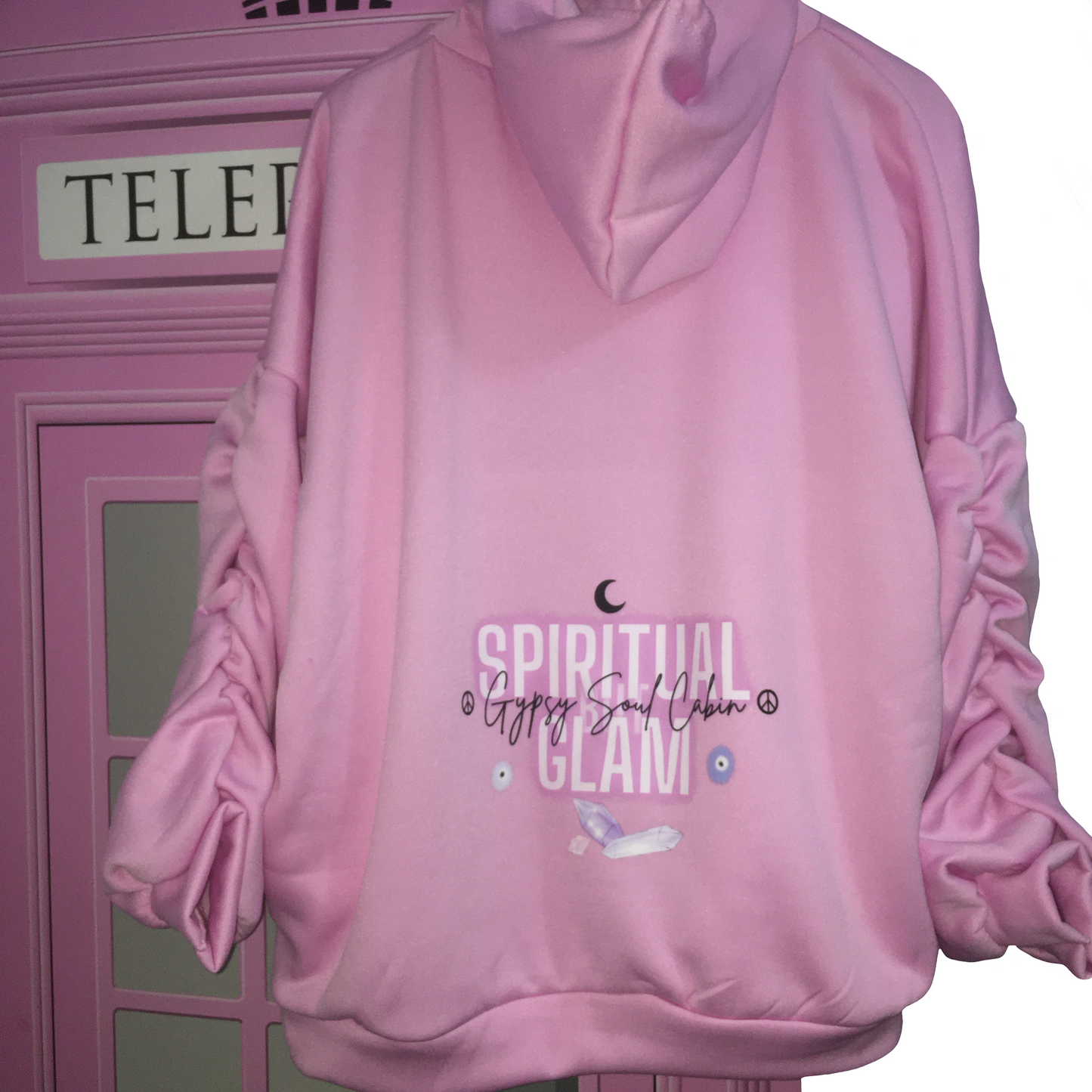 A pink hoodie with 'SPIRITUAL BUT GLAM' printed on the back, featuring a crescent moon, purple crystals, and an evil eye graphic. The hoodie has ruched sleeves and is displayed against a pink phone booth backdrop.