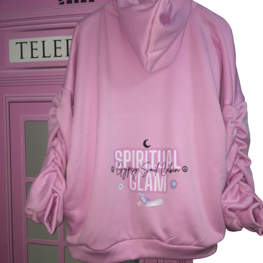 A pink hoodie with 'SPIRITUAL BUT GLAM' printed on the back, featuring a crescent moon, purple crystals, and an evil eye graphic. The hoodie has ruched sleeves and is displayed against a pink phone booth backdrop.