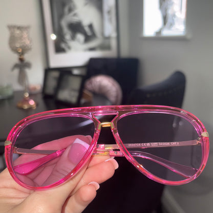 A hand holding a pair of aviator-style sunglasses with a translucent hot pink frame and dark lenses, set against a blurred indoor background.