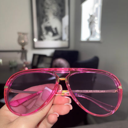 A hand holding a pair of oversized aviator-style sunglasses with translucent hot pink frames and dark tinted lenses in a blurred indoor setting.