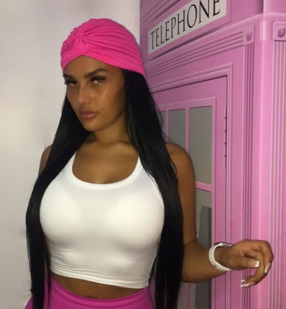 Front view of a woman wearing a pink bohemian gypsy head wrap, white crop top, and pink trousers, standing against a pink background with aesthetic wardrobe details visible.