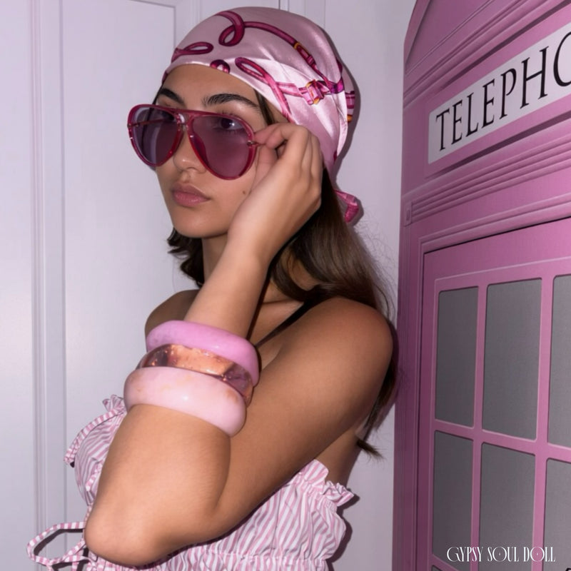 A woman wearing a pink evil eye patterned silk boho head scarf, pink aviator sunglasses, and a pink and white striped top, posing next to a pink telephone booth prop by Gypsy Soul Doll.