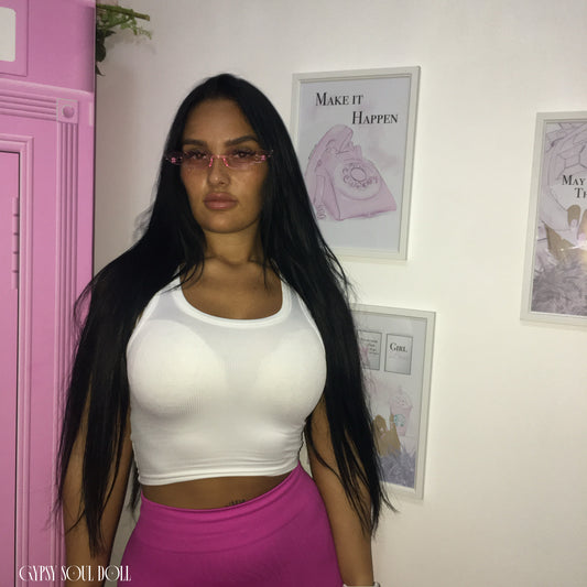 A Gypsy Soul Doll model with long black hair and pink-framed fashion glasses stands indoors wearing a white crop top and a bright pink skirt. The background shows a white wall with framed art and a pink phone booth prop on the left.