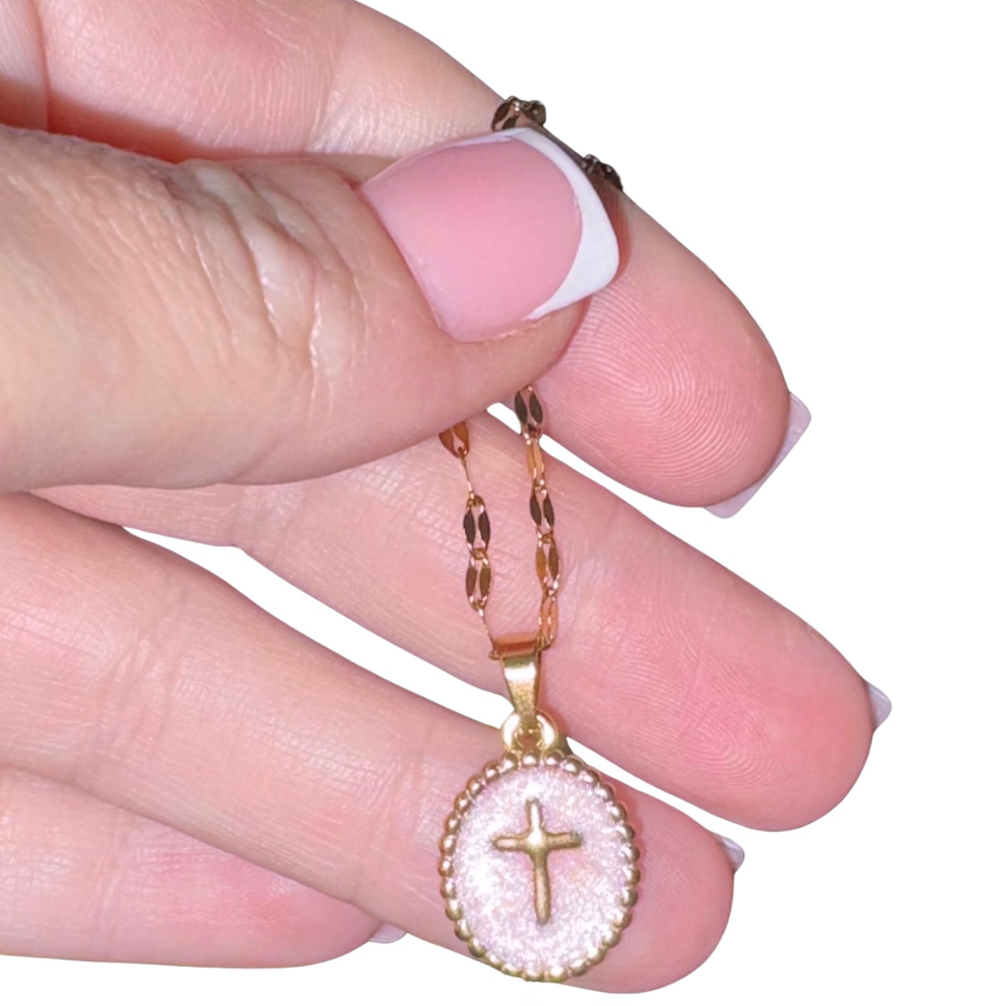 A close-up of a hand holding a gold chain necklace with a round pendant featuring a gold cross on a textured pale pink background.