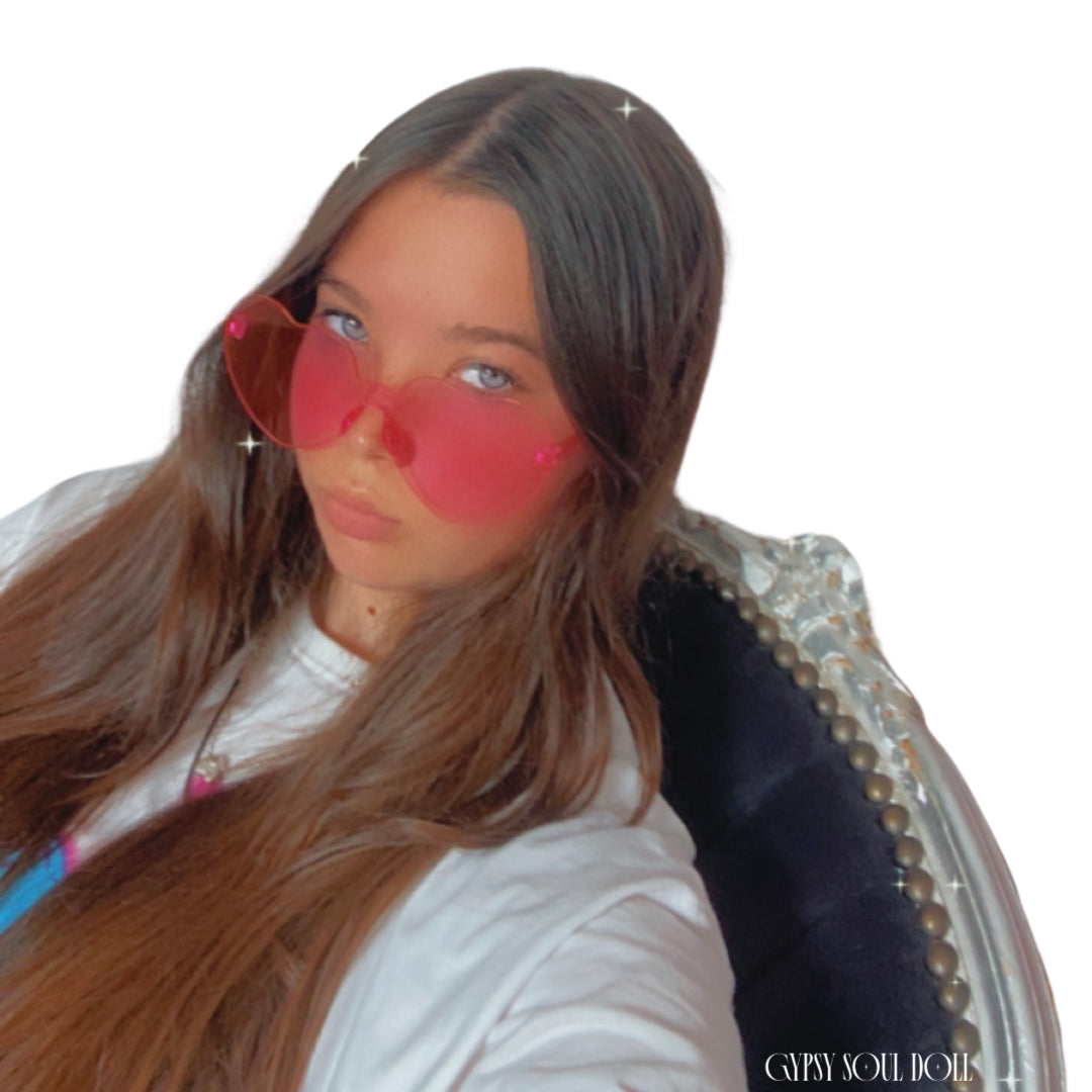 A girl with long brown hair wearing translucent pink heart-shaped fashion glasses and a white t-shirt, sitting in a dark velvet French chair with gold nailhead trim against a white background.