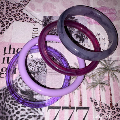 Four stacked translucent bangles in different shades of purple and lavender, photographed on a patterned pink and white surface by Gypsy Soul Doll.