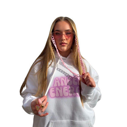 A young woman with long blonde hair and pink chain-link glasses is wearing a white hoodie with purple lettering that reads 'ANGEL ENERGY'. With matching pink accessories against a solid white backgroour.
