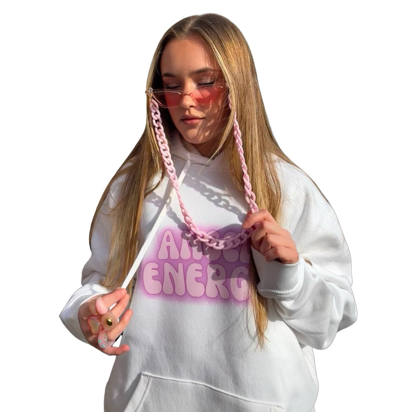 A young model with long blonde hair wearing a white hoodie with a pink graphic that reads 'ANGEL ENERGY' with a pink fashion chain lanyard and matching accessories, posed against a solid white background.