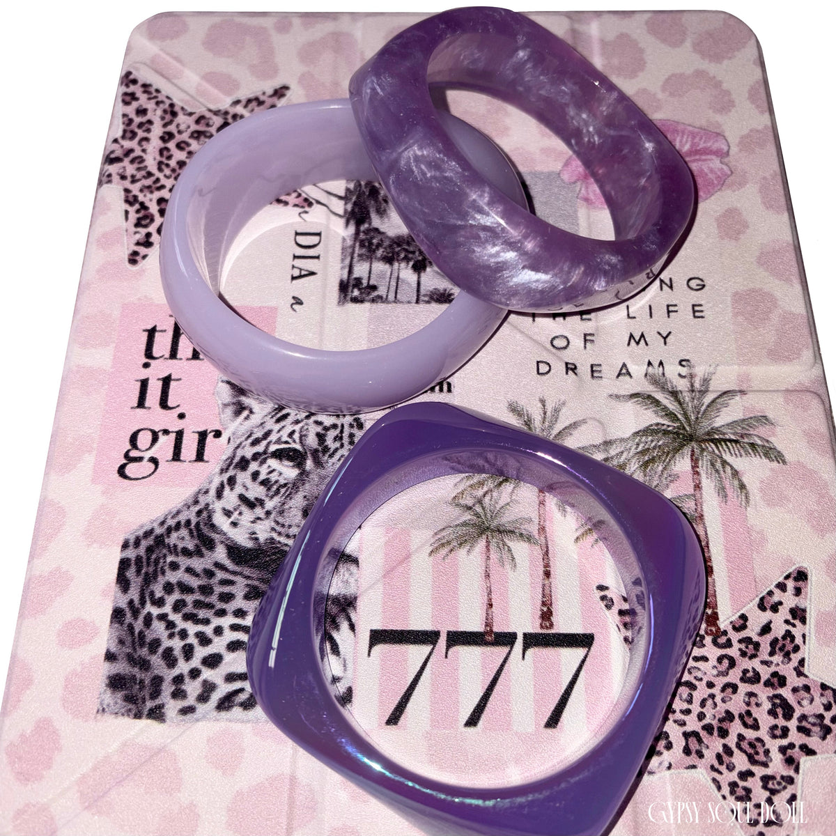 Flat lay view of the three-piece purple resin bangle collection arranged over a pink leopard print journal styled with the number 777 and It-girl text.