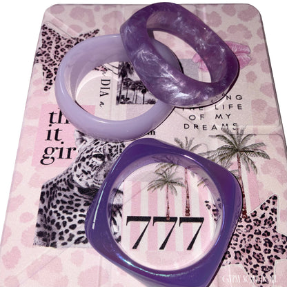 Flat lay view of the three-piece purple resin bangle collection arranged over a pink leopard print journal styled with the number 777 and It-girl text.