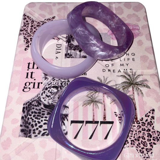 Flat lay view of the three-piece purple resin bangle collection arranged over a pink leopard print journal styled with the number 777 and It-girl text.