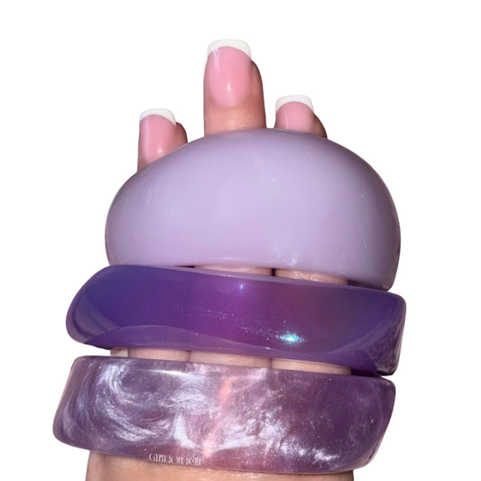 Close-up hand view showcasing a three-piece purple candy acrylic bangle stack featuring a thick pastel lavender dome bangle, a translucent dark purple bangle, and a metallic marbled violet bangle.