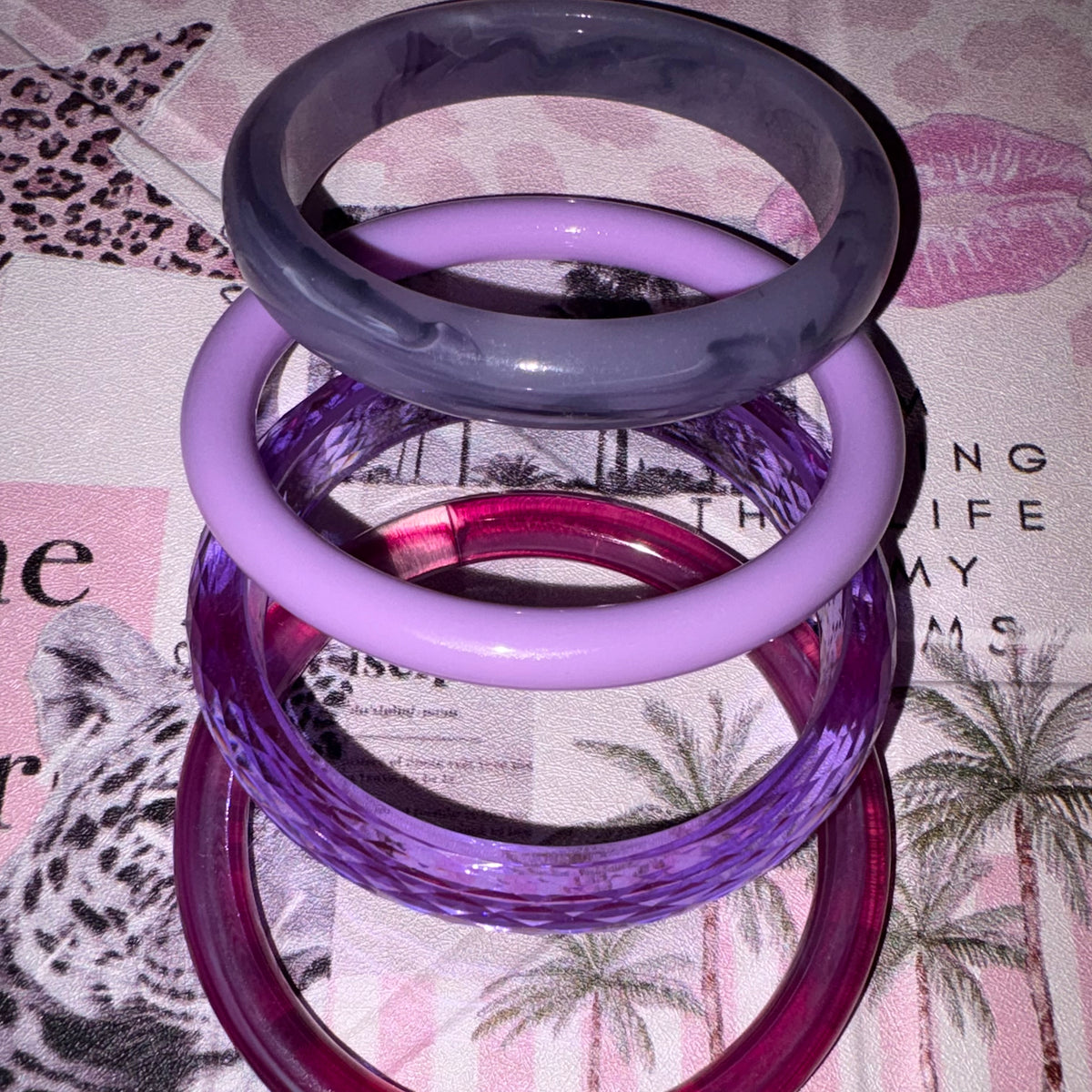 Four stacked translucent bangles in different shades of purple and lavender, photographed on a patterned pink and white surface.
