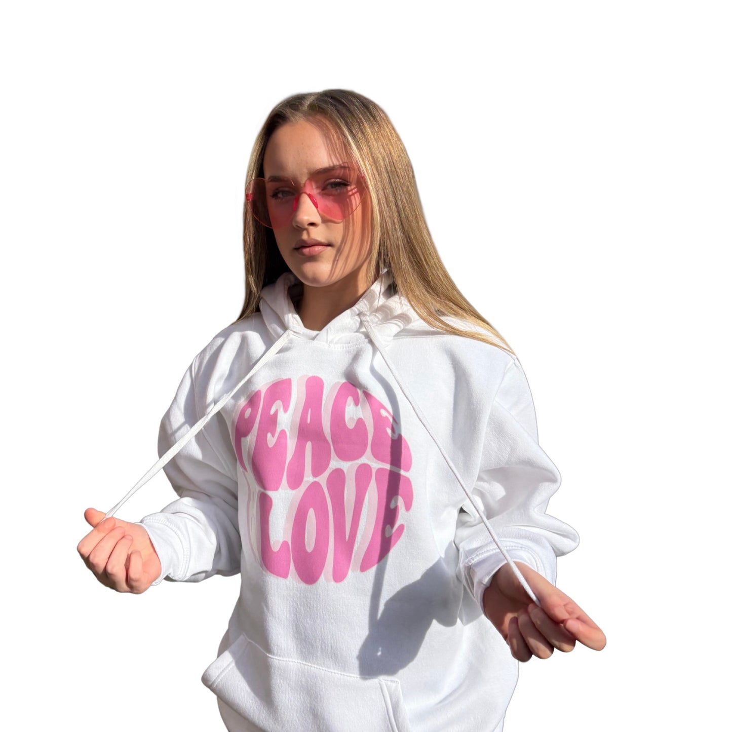 A young model with long blonde hair and pink-tinted fashion glasses wears a white hoodie with a bold pink 'PEACE LOVE' graphic on the front, holding the drawstrings against a solid white background.