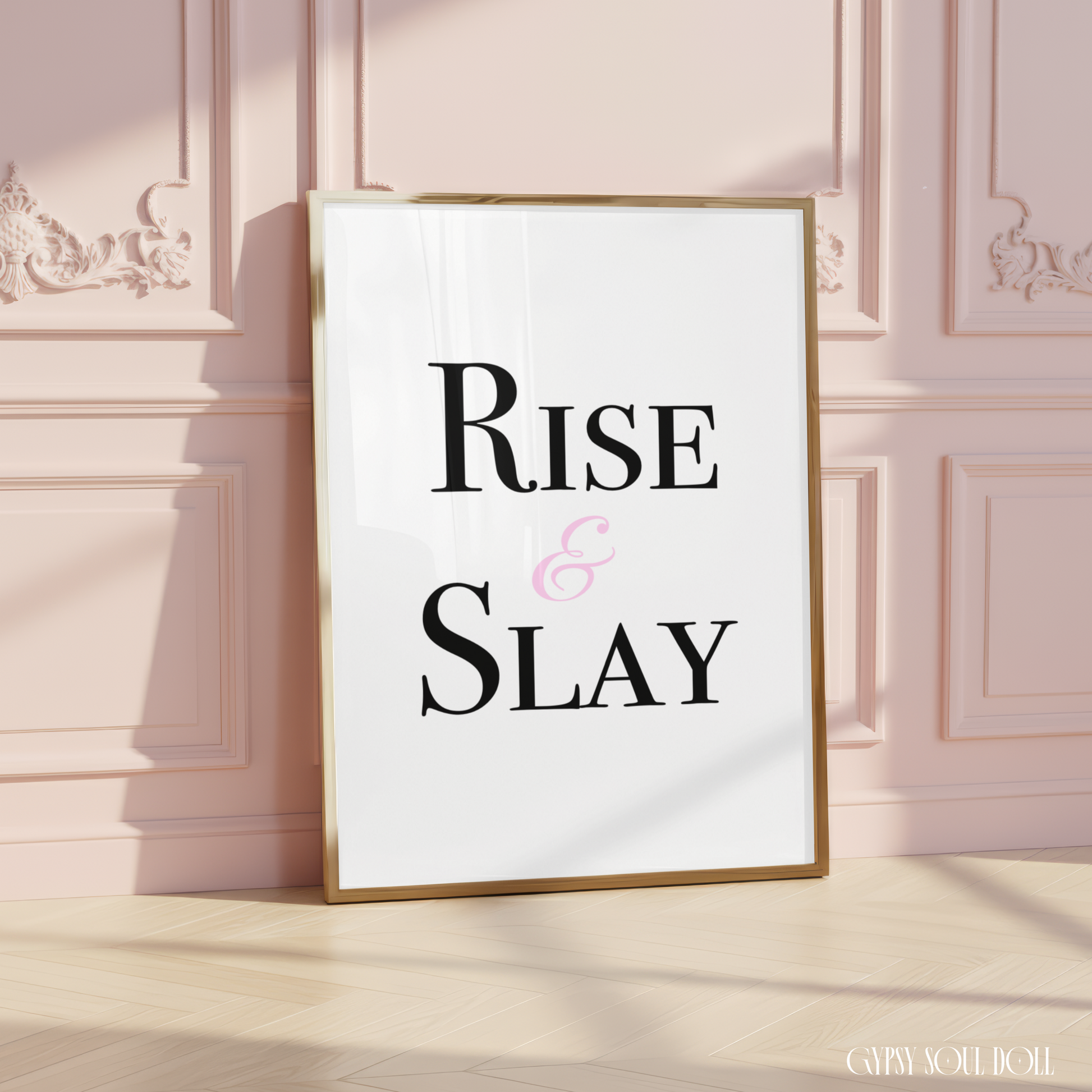 A framed typographic wall art print reading “RISE & SLAY” in black serif text with a pink ampersand on a white background, leaning against a pale pink paneled wall on a light wood floor.