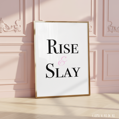A framed typographic wall art print reading “RISE & SLAY” in black serif text with a pink ampersand on a white background, leaning against a pale pink paneled wall on a light wood floor.