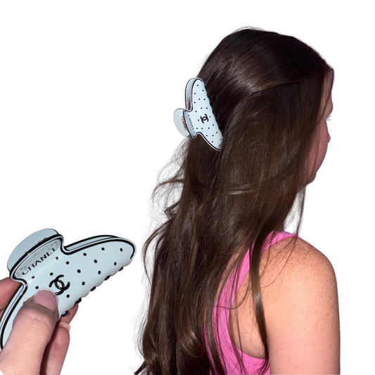 A Chanel hair claw clip with a white and black polka dot pattern and the iconic interlocking CC logo, shown both being held by a hand and worn by a model with long brown hair.