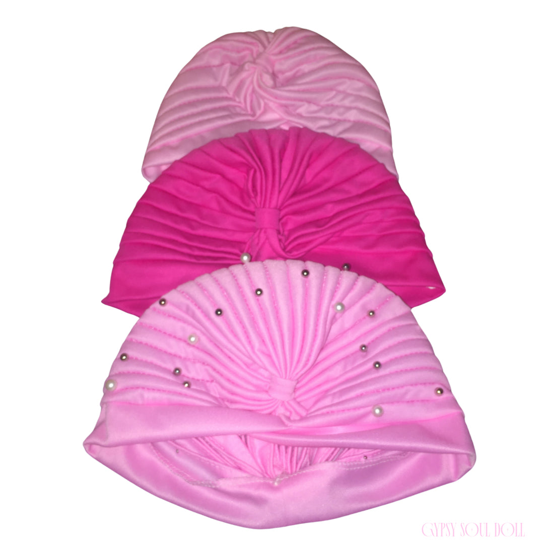A vertical stack layout showcasing three variations of the Gypsy Soul Doll Sahara pleated head wrap turbans: a pastel baby pink version, a bright hot pink style, and a baby pink variant embellished with scattered silver pearls and metallic studs against a plain white background.