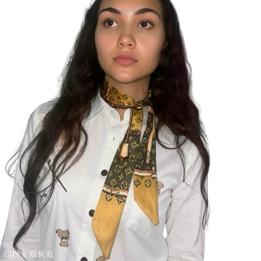 Model showcasing the Gypsy Soul Doll khaki and gold monogram skinny satin scarf wrapped elegantly as a multi-way neck tie choker, styled next to a pink telephone box.